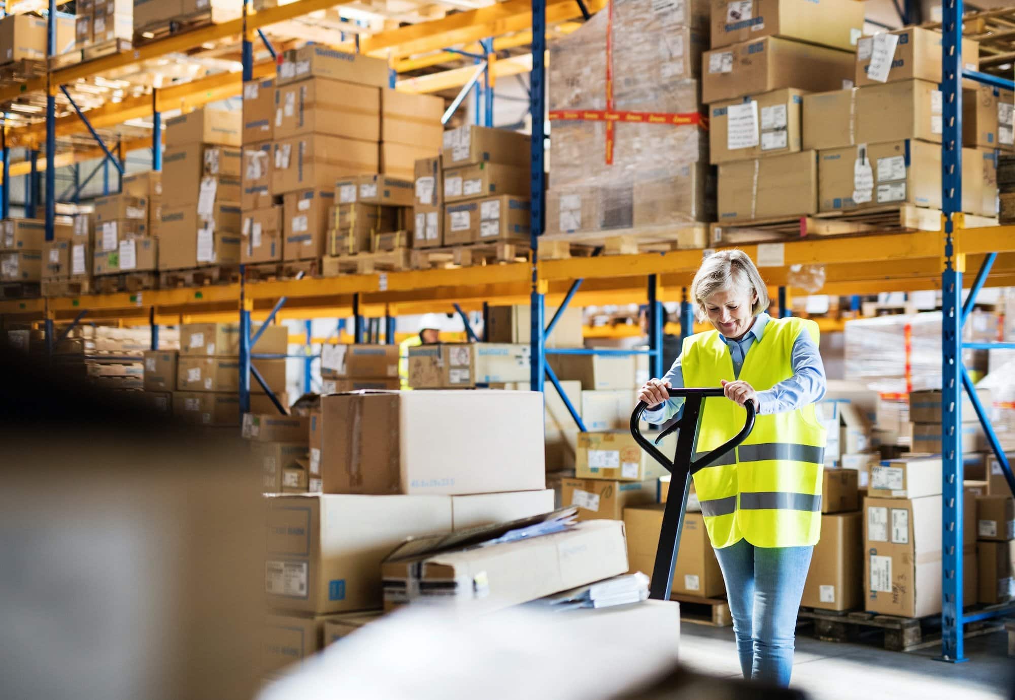 A senior woman warehouse worker pulling a pallet truck with boxes.